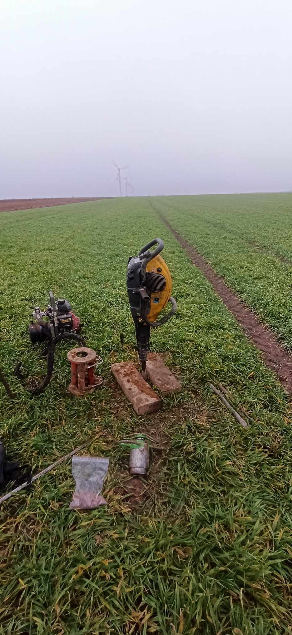 Praca geologiczna w terenie: wiertnica spalinowa wbija się w grunt na polu, w tle widoczne turbiny wiatrowe we mgle.