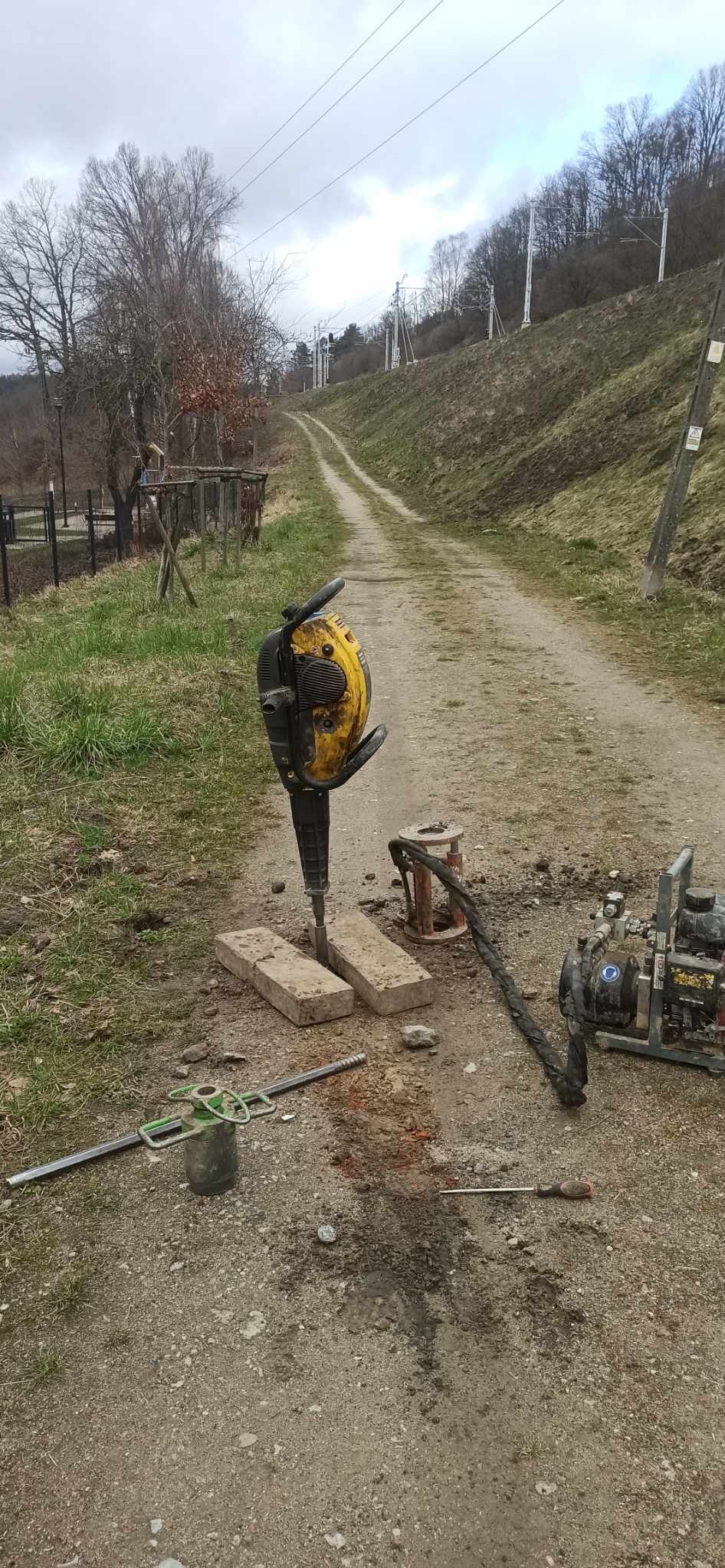 Żółto-czarny młot udarowy wbity w dwie betonowe płyty na polnej drodze, w tle linia kolejowa na zboczu.