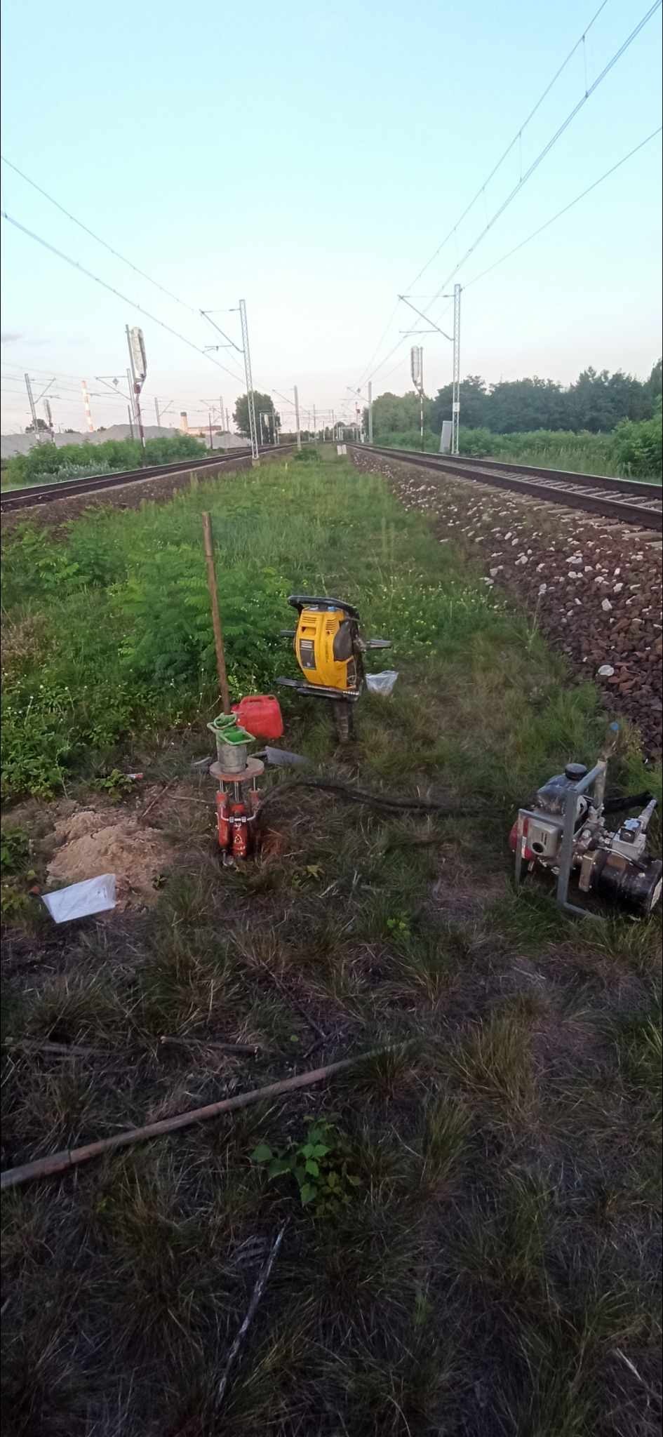 Prace w terenie przy torach kolejowych z użyciem wiertnicy geologicznej i pompy wodnej. Widoczne narzędzia i próbki gruntu.