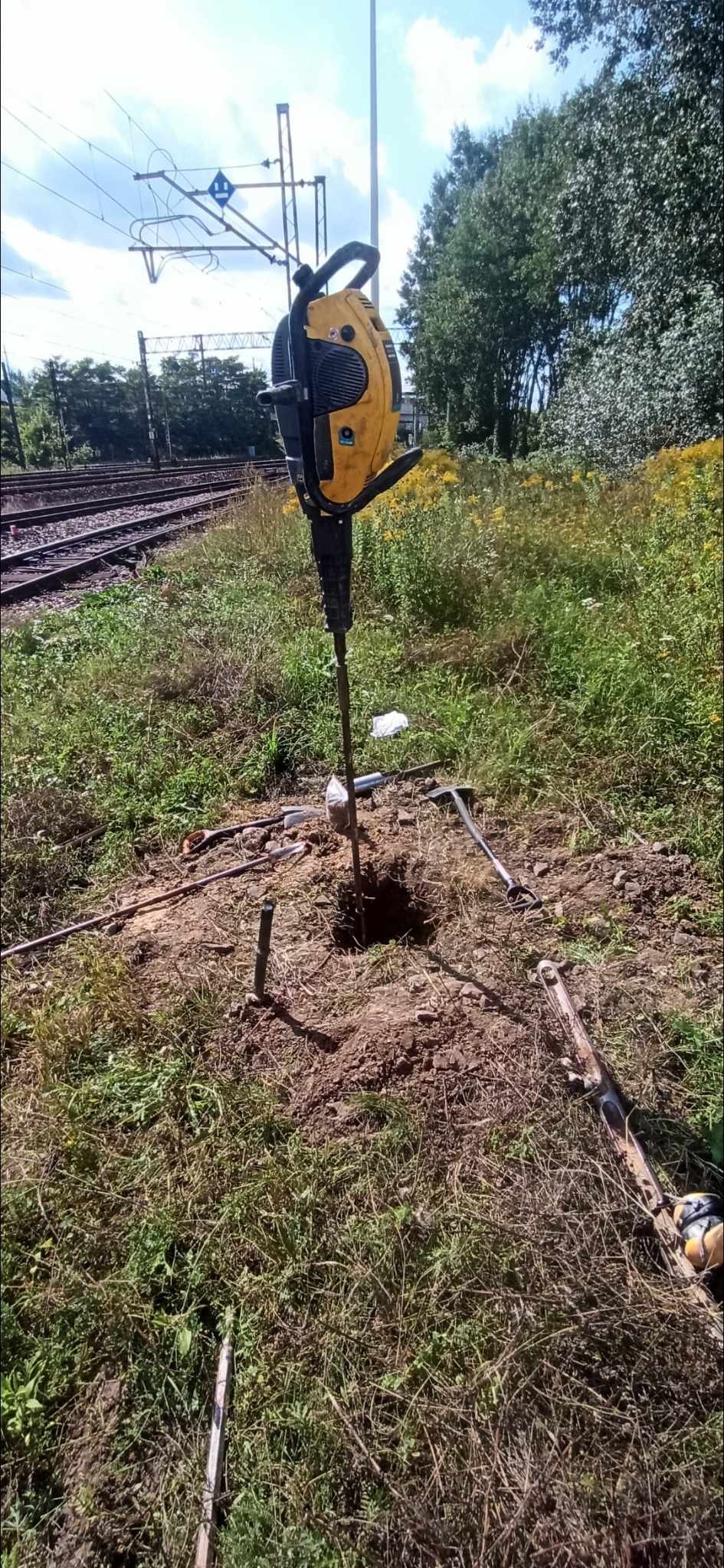 Żółty wiertniczy młot udarowy wbija się w ziemię na zewnątrz, obok torów kolejowych, z rozrzuconymi narzędziami.