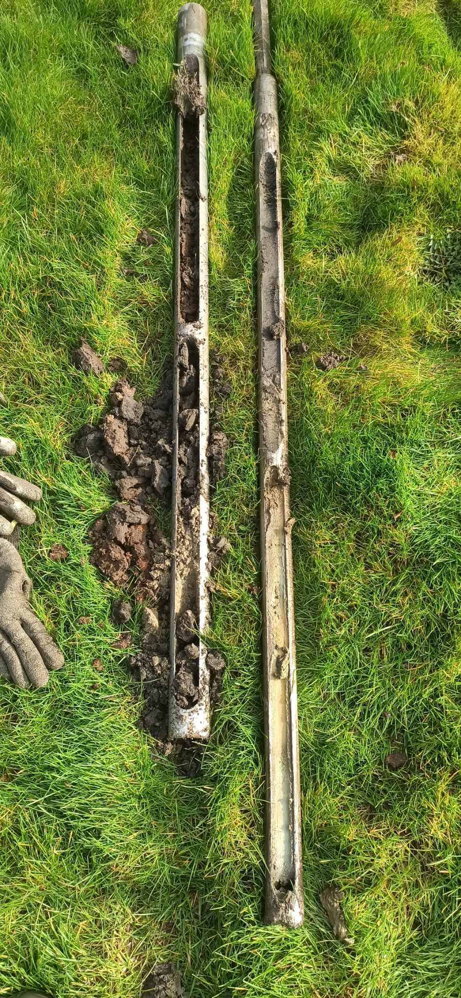 Dwa rdzenie glebowe w metalowych rurach leżą na trawie, jeden z widocznymi bryłami ziemi, drugi z bardziej jednolitą próbką, obok leżą rękawice robocze.