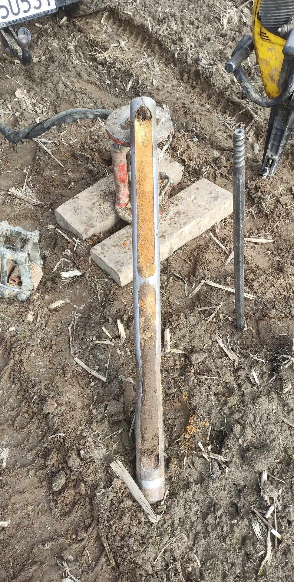 Pobieranie próbki gruntu za pomocą świdra rdzeniowego na placu budowy, widoczna maszyna wibracyjna i fragment tablicy rejestracyjnej pojazdu.