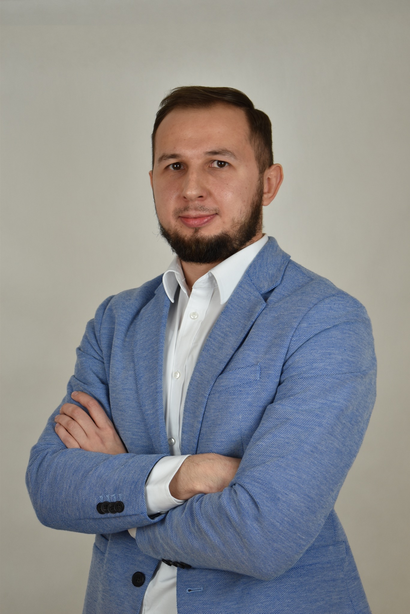 Portret mężczyzny w błękitnej marynarce i białej koszuli, z brodą i rękami skrzyżowanymi na piersiach, na neutralnym tle.