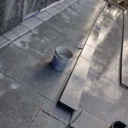 Układanie szarej kostki brukowej na tarasie, widoczny kubeł z zaprawą i narzędzia, w tle folia ochronna i rusztowanie.