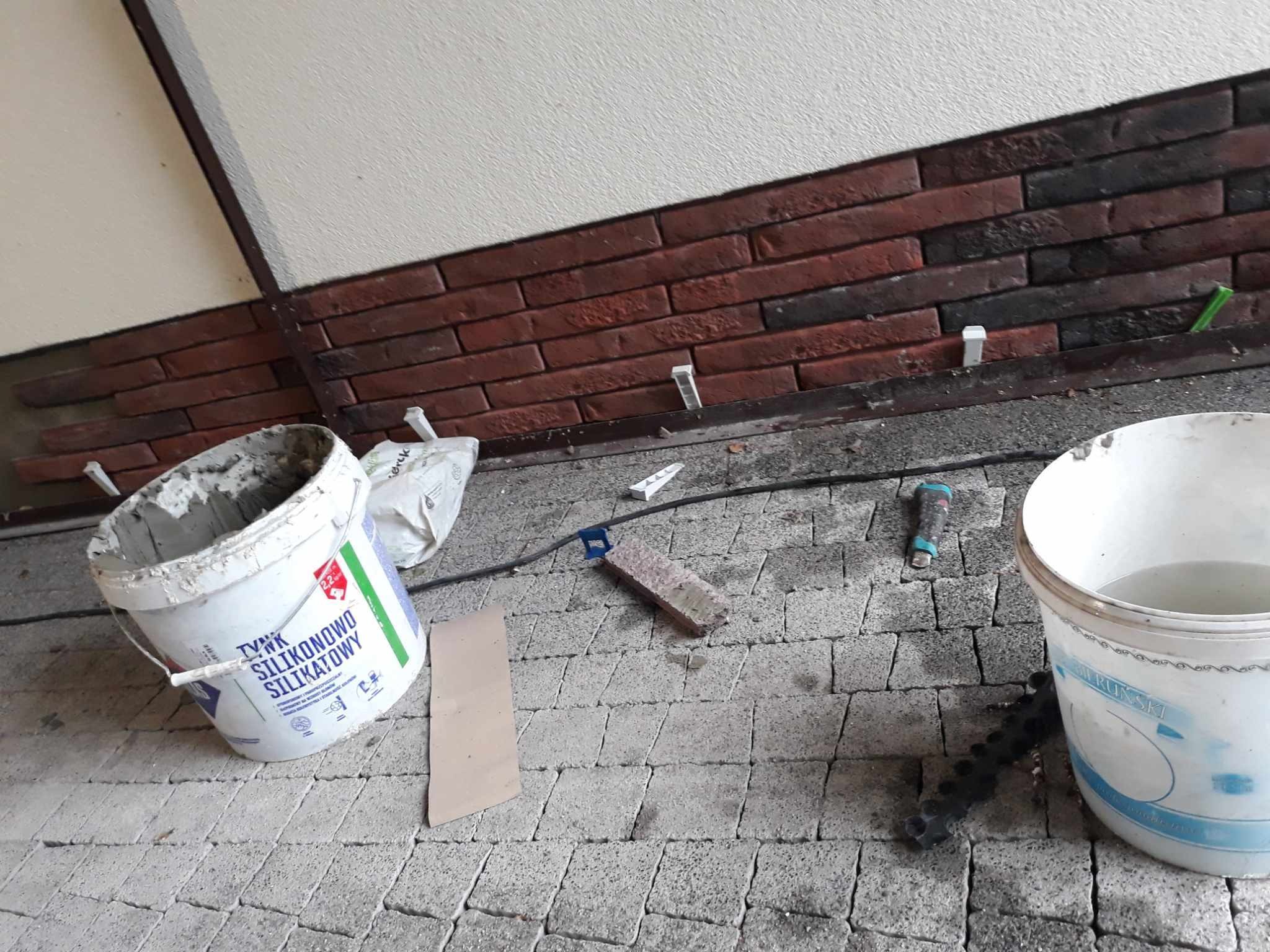 Fragment ściany z klinkieru w trakcie montażu, widoczne wiaderka z zaprawą, narzędzia i elementy mocujące na kostce brukowej.