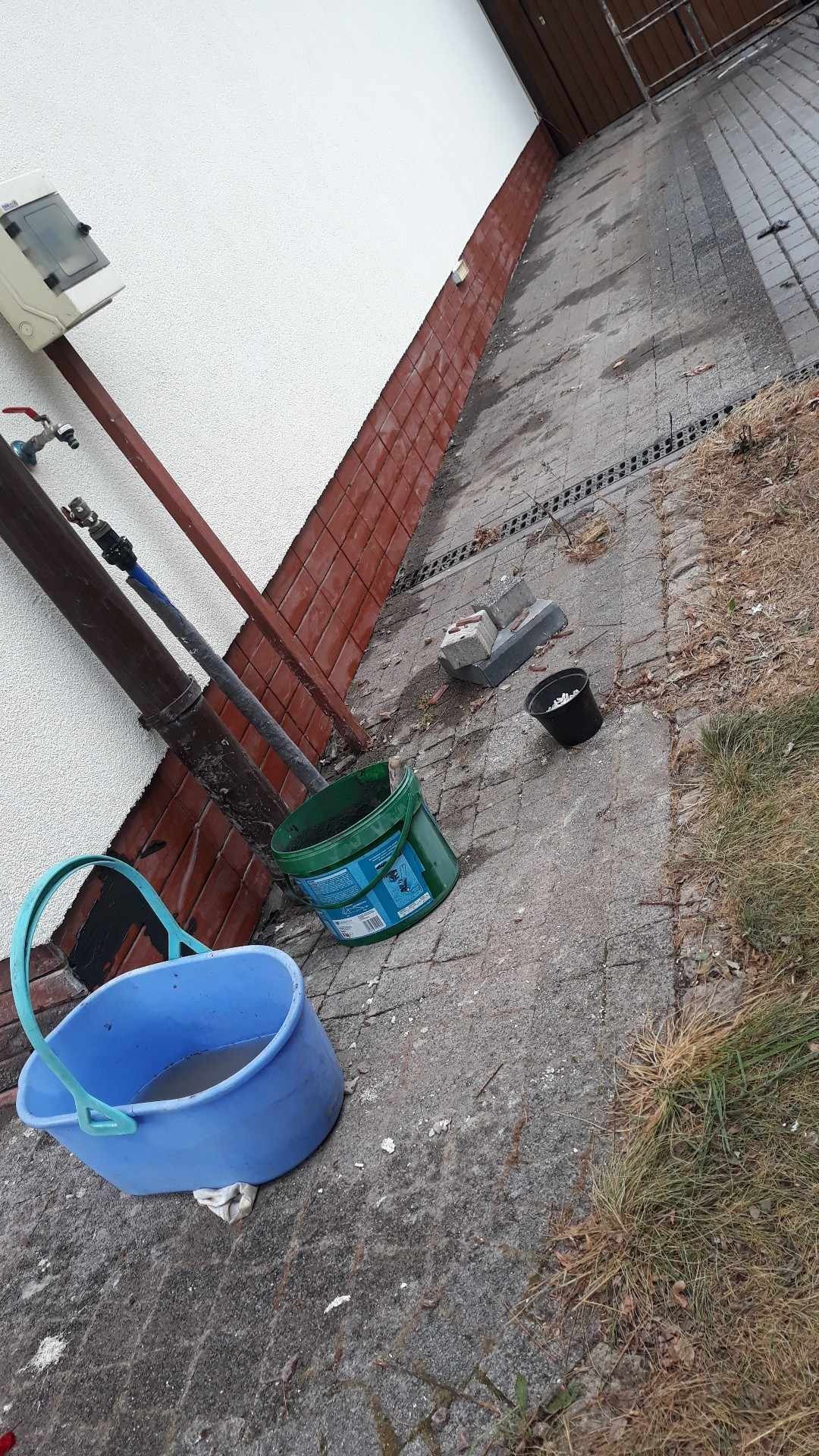 Dwa wiadra z farbą i gruzem stojące na betonowej kostce brukowej obok ściany domu z rurą spustową i licznikiem gazu.