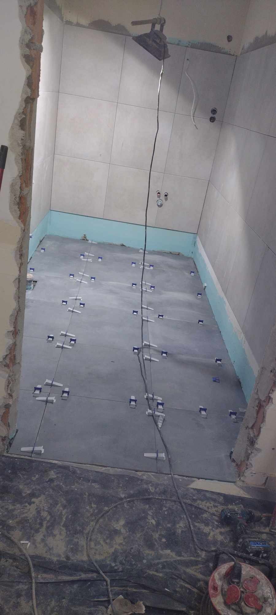 Świeżo ułożone szare płytki podłogowe w łazience, z widocznymi niebieskimi klipsami poziomującymi, kabel zwisający z sufitu, fragment ściany z uszkodzonym tynkiem po lewej stronie oraz narzędzia...