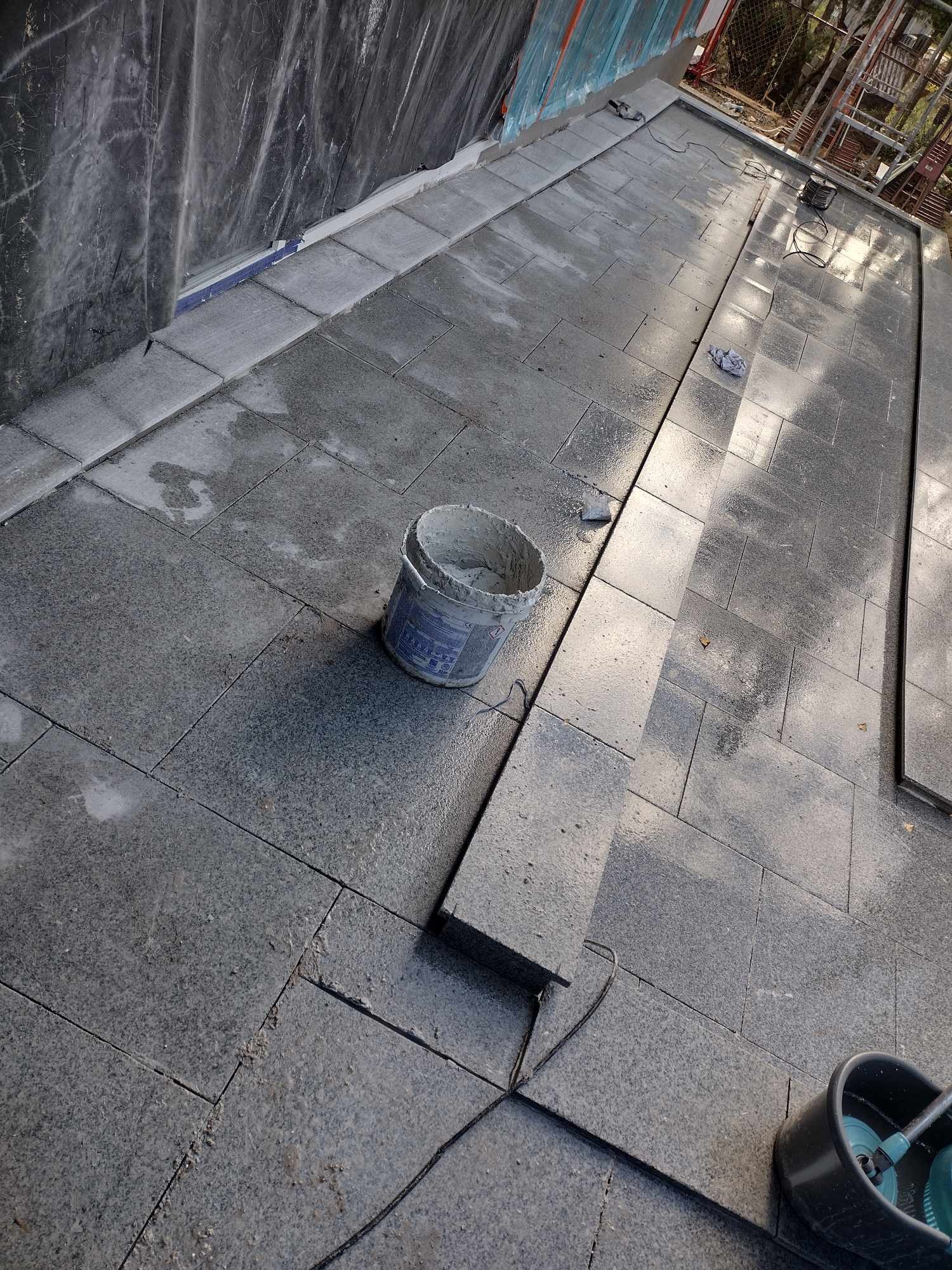 Układanie szarej kostki brukowej na tarasie, widoczny kubeł z zaprawą i narzędzia, w tle folia ochronna i rusztowanie.