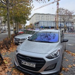 Kml_wyklejanki - Srebrny Hyundai i30 z przyciemnioną szybą czołową, zaparkowany na brukowanej ulicy, zasypanej jesiennymi liśćmi. W tle budynek i żurawie budowlane.