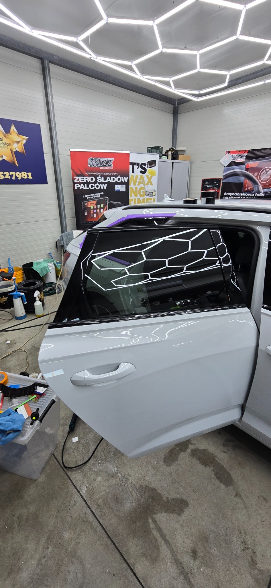 Biały samochód z otwartymi drzwiami w warsztacie, na szybie wzór plastra miodu. Widoczne narzędzia i reklamy firmowe w tle. Oświetlenie sufitowe w kształcie heksagonów.