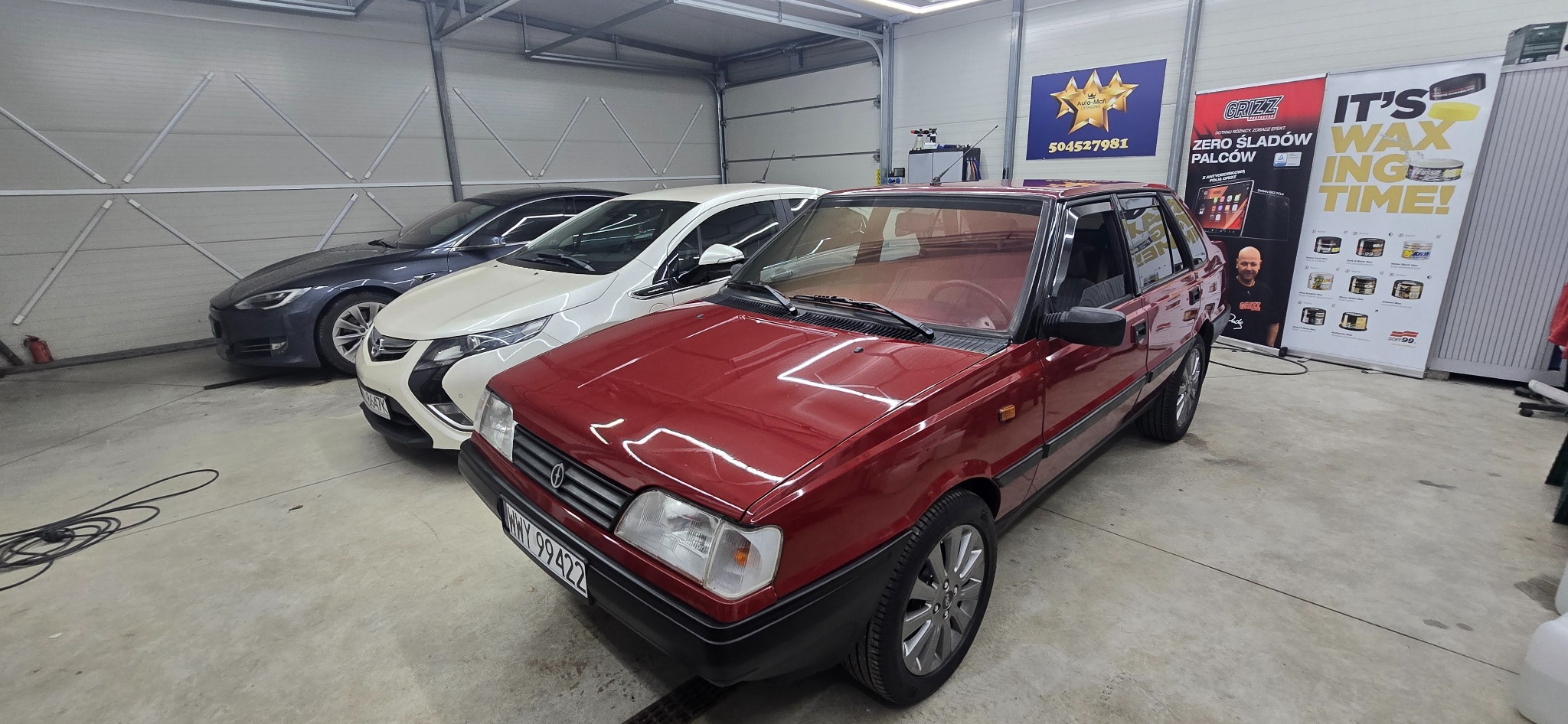 Bordowy Polonez Caro po detailingu, zaparkowany w garażu obok Tesli i Opla. Błyszcząca karoseria, widoczne reklamy środków do pielęgnacji aut.
