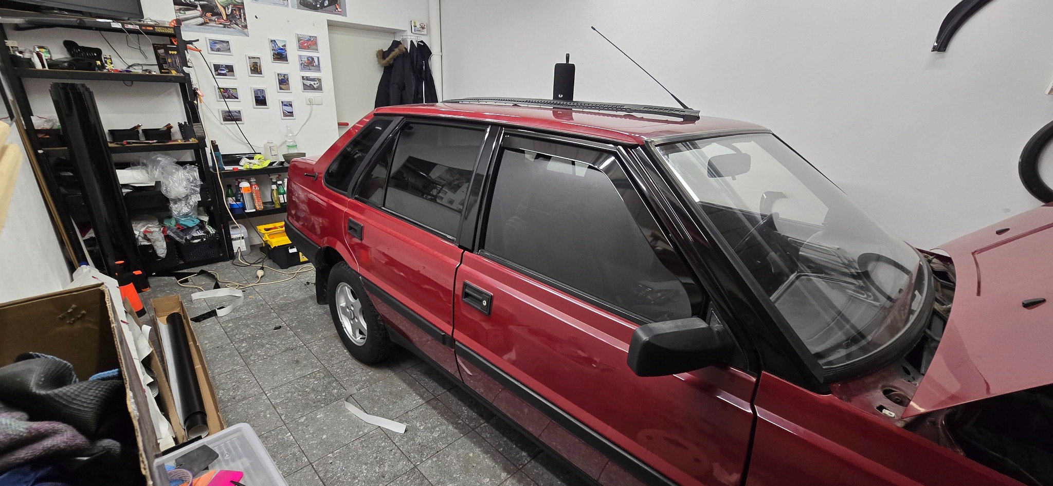 Czerwony samochód z przyciemnionymi szybami bocznymi, zaparkowany w warsztacie. Widoczne otwarte drzwi i podniesiona maska. Na półkach narzędzia i akcesoria.