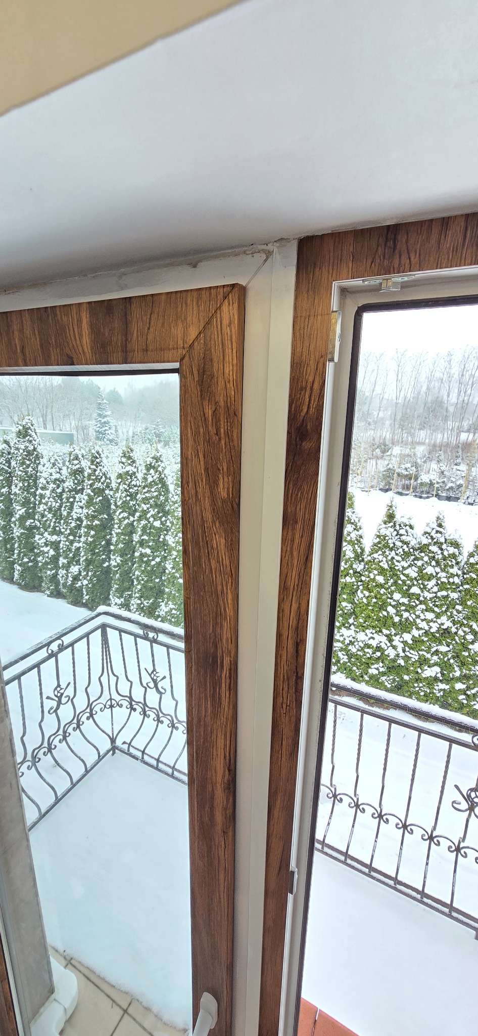 Okleina imitująca drewno na ramie okiennej z widokiem na zasypany śniegiem balkon i drzewa. Dekoracja okna z bliska w zimowej scenerii.