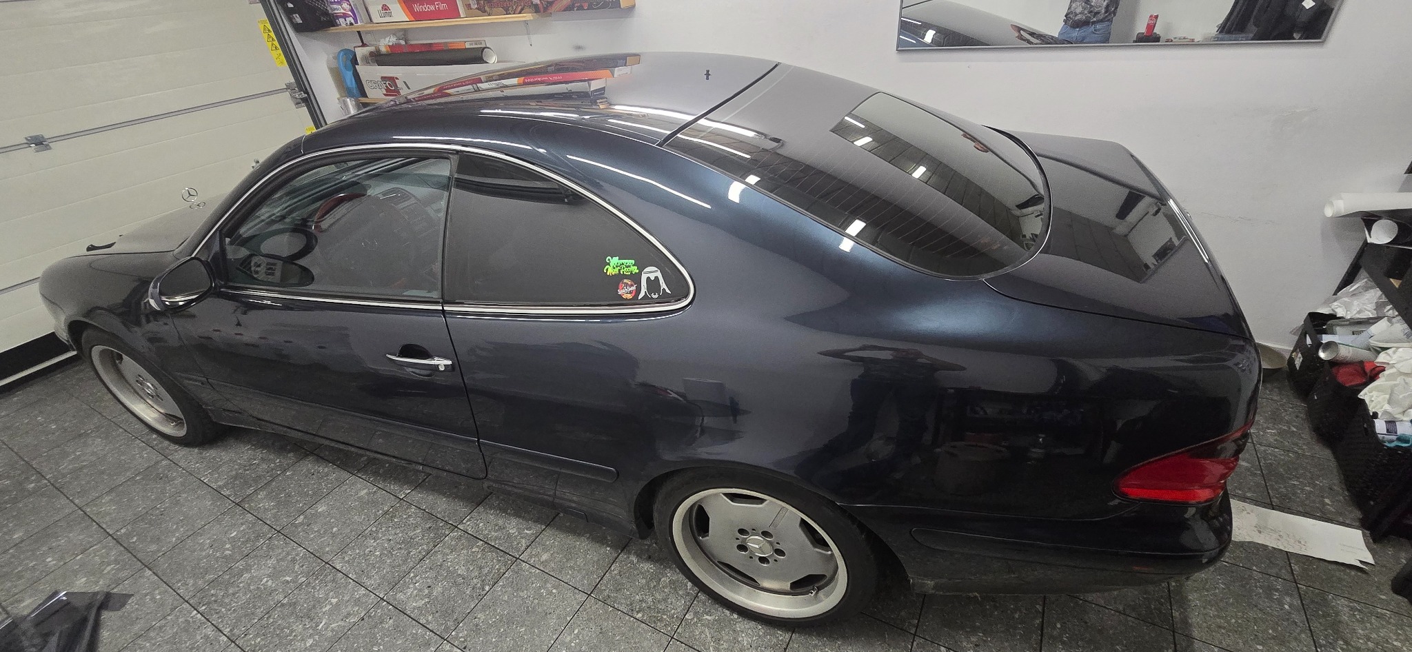 Ciemny Mercedes CLK z przyciemnionymi szybami, widoczne naklejki na tylnej szybie. Auto stoi w garażu na szarej podłodze z płytek.