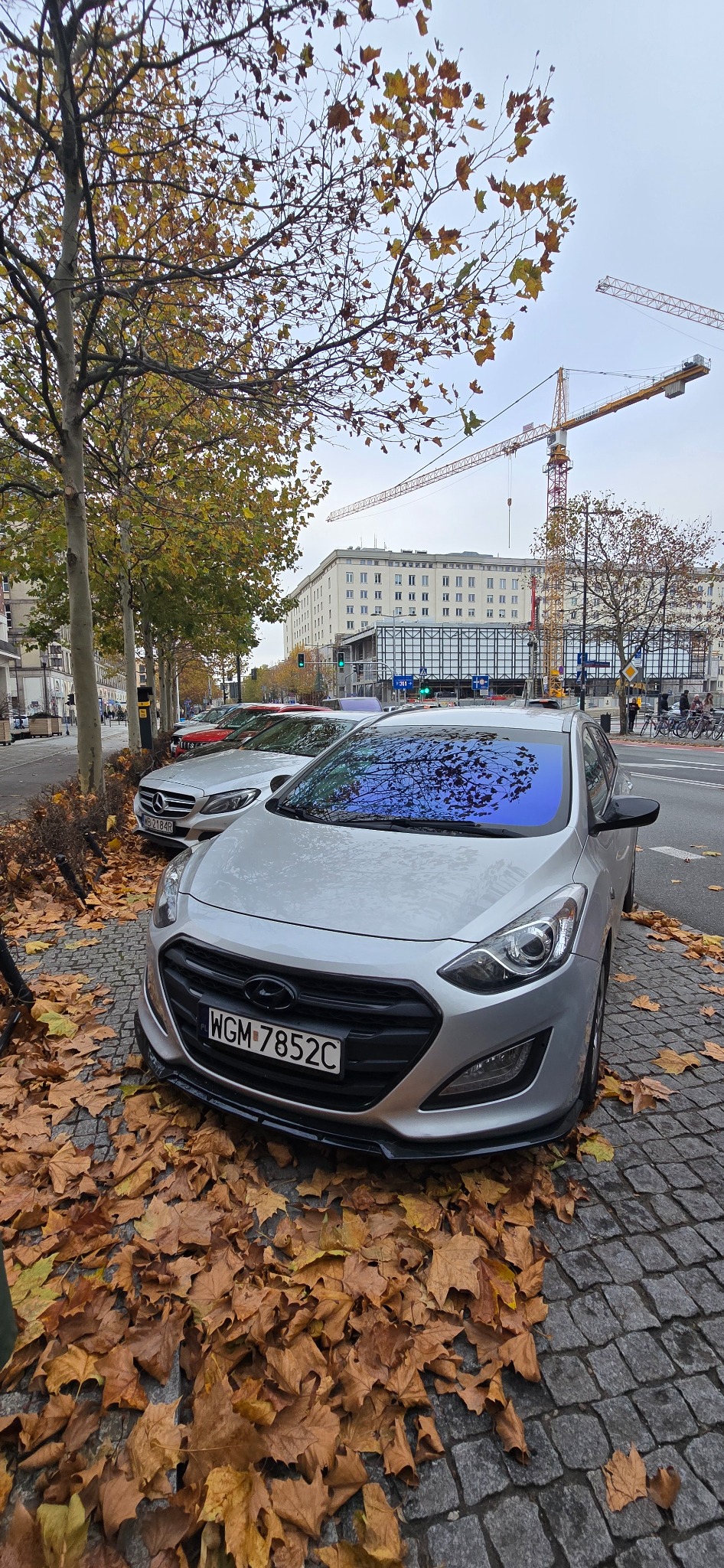 Srebrny Hyundai i30 z przyciemnioną szybą czołową, zaparkowany na brukowanej ulicy, zasypanej jesiennymi liśćmi. W tle budynek i żurawie budowlane.