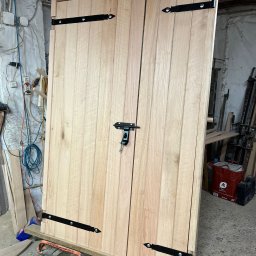 ERGO Mariusz Czajka - Surowe dwuskrzydłowe drewniane drzwi z czarnymi zawiasami i zasuwką, umieszczone na wózku transportowym w warsztacie.