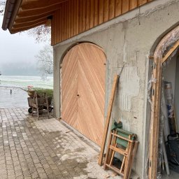 ERGO Mariusz Czajka - Nowa, drewniana brama garażowa z ukośnym wzorem desek, zamontowana w surowym, betonowym murze budynku w trakcie budowy, widok z zewnątrz na podjazd i zimowy krajobraz.