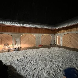 ERGO Mariusz Czajka - Nowo wybudowany garaż z czterema drewnianymi bramami łukowymi, widok nocą, śnieg na podłożu, oświetlenie wewnętrzne bram.