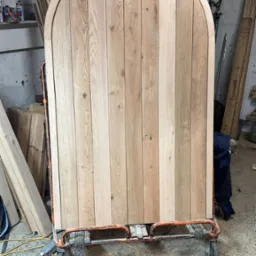 Duży, zaokrąglony panel z jasnego drewna na wózku transportowym w warsztacie, widoczne sęki i usłojenie drewna.