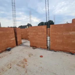 Wnętrze w trakcie budowy domu z czerwonej cegły, widoczne ściany z otworami na okna i drzwi, zbrojenie pionowe w narożnikach, betonowa podłoga z resztkami zaprawy i narzędzia budowlane w rogu.