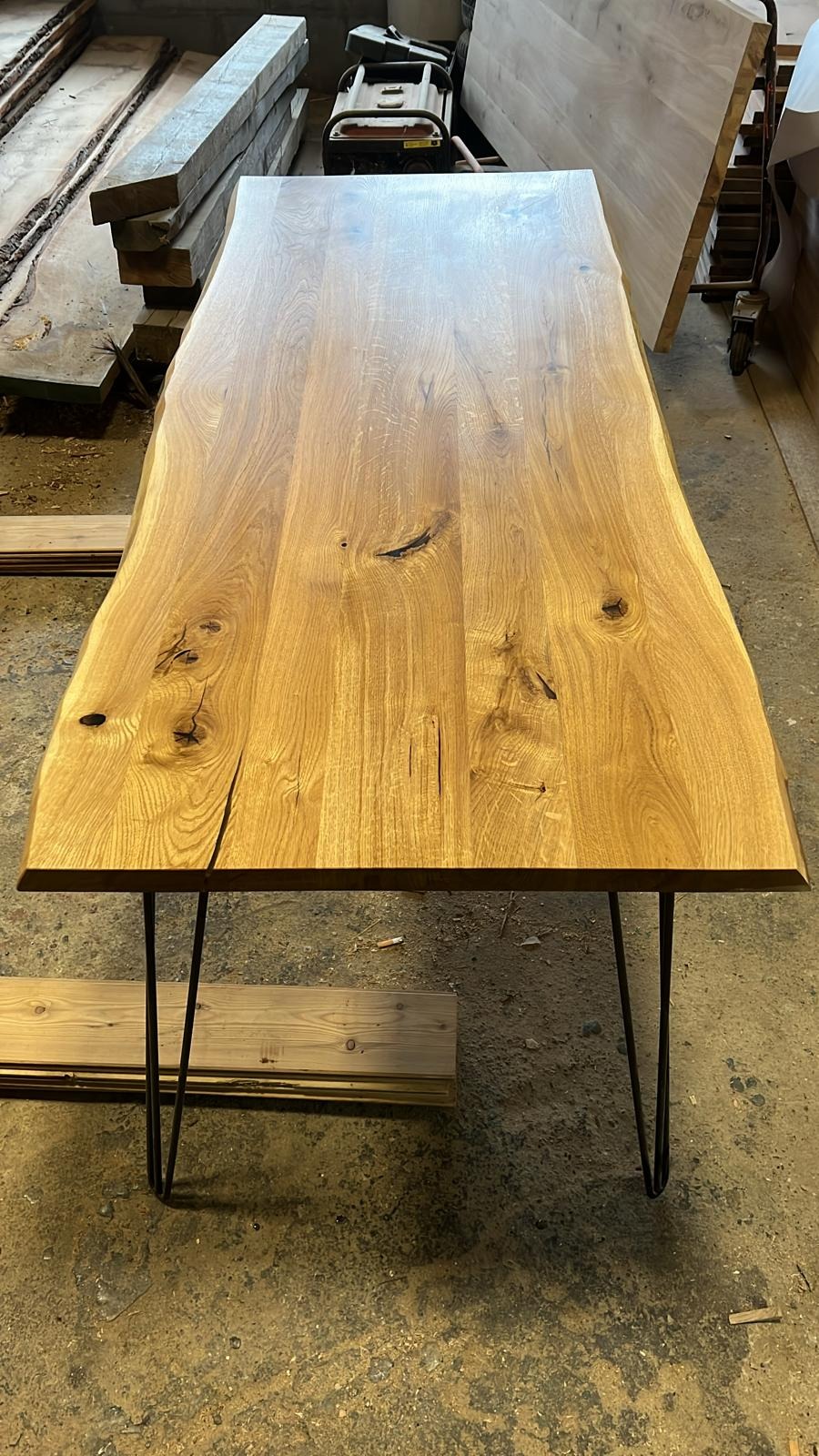 Surowy blat z litego drewna z widocznymi sękami i pęknięciami, osadzony na czarnych, metalowych nogach typu hairpin, w tle składowane deski.