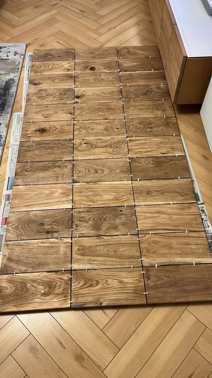 Ułożone na podłodze drewniane płytki imitujące deski, z krzyżykami dystansowymi, przygotowane do montażu blatu kuchennego na tle parkietu w jodełkę.
