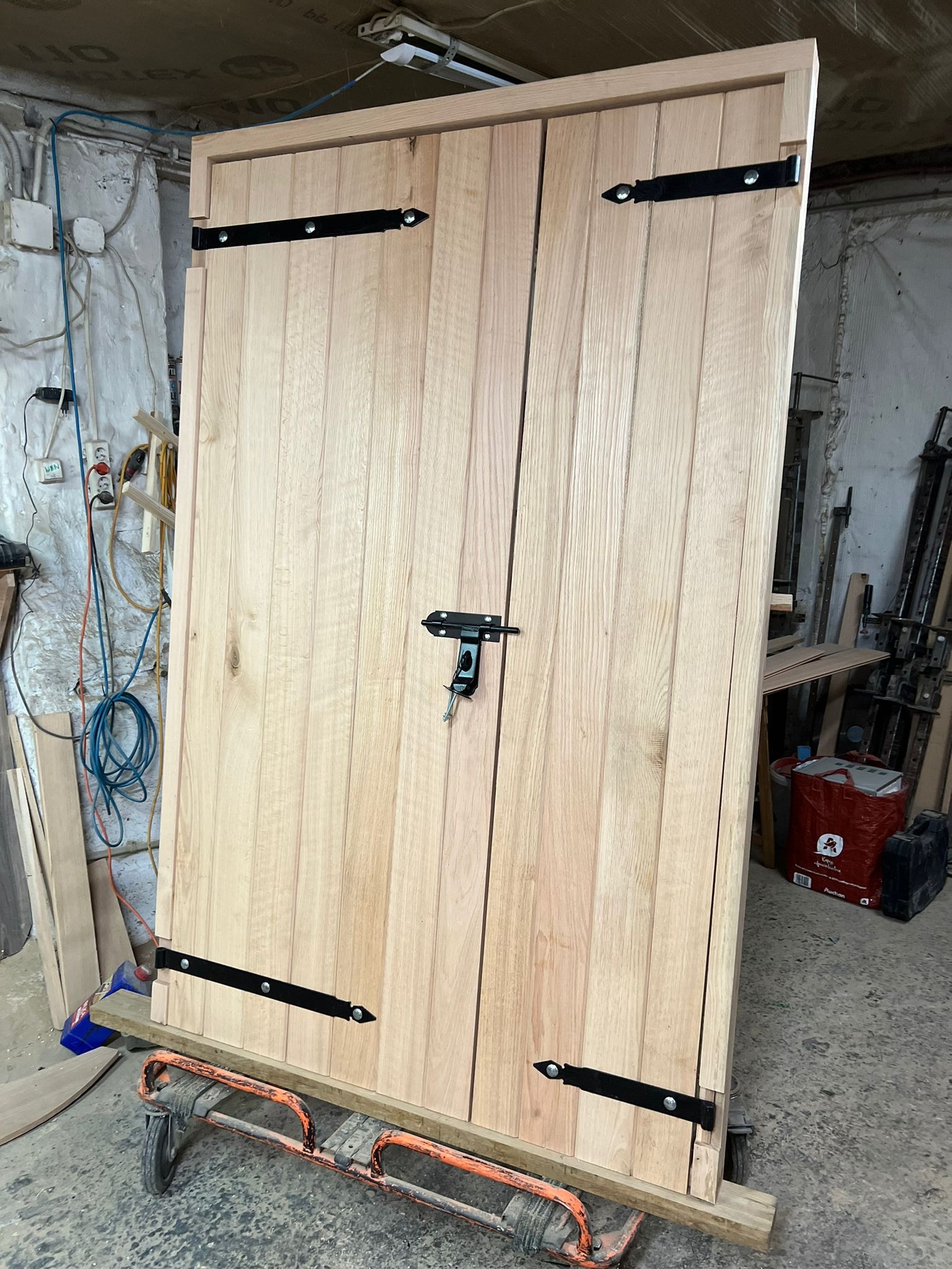 Surowe dwuskrzydłowe drewniane drzwi z czarnymi zawiasami i zasuwką, umieszczone na wózku transportowym w warsztacie.