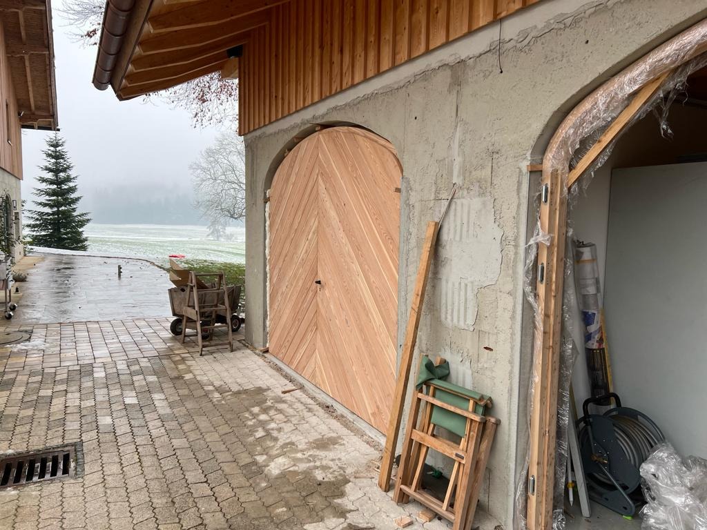 Nowa, drewniana brama garażowa z ukośnym wzorem desek, zamontowana w surowym, betonowym murze budynku w trakcie budowy, widok z zewnątrz na podjazd i zimowy krajobraz.