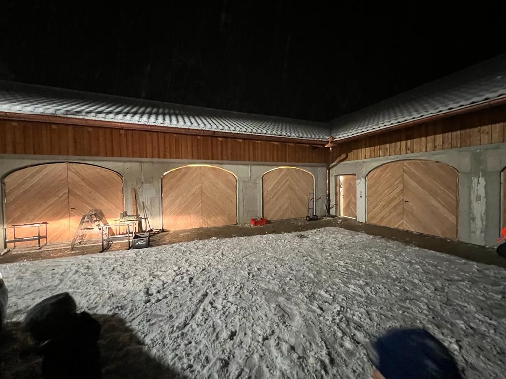Nowo wybudowany garaż z czterema drewnianymi bramami łukowymi, widok nocą, śnieg na podłożu, oświetlenie wewnętrzne bram.