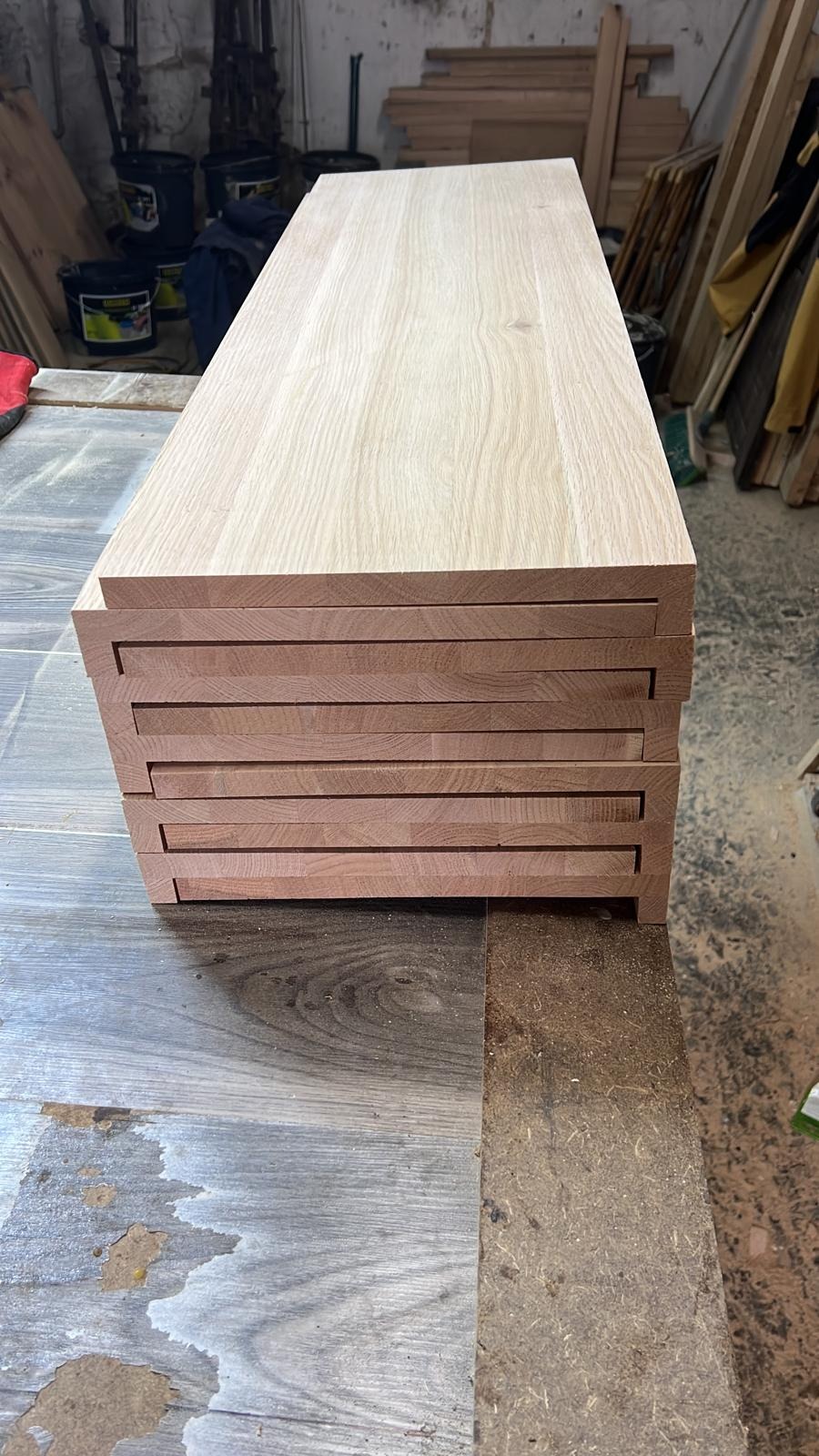 Stos surowych, przygotowanych do montażu blatów z jasnego drewna ułożonych jeden na drugim w warsztacie, widoczne frezy na krawędziach.