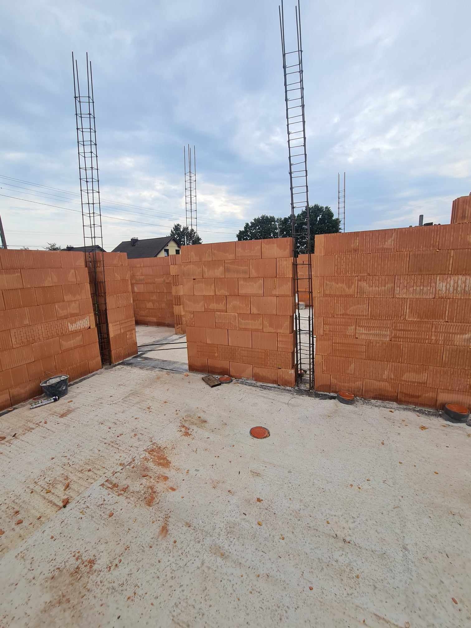Wnętrze w trakcie budowy domu z czerwonej cegły, widoczne ściany z otworami na okna i drzwi, zbrojenie pionowe w narożnikach, betonowa podłoga z resztkami zaprawy i narzędzia budowlane w rogu.
