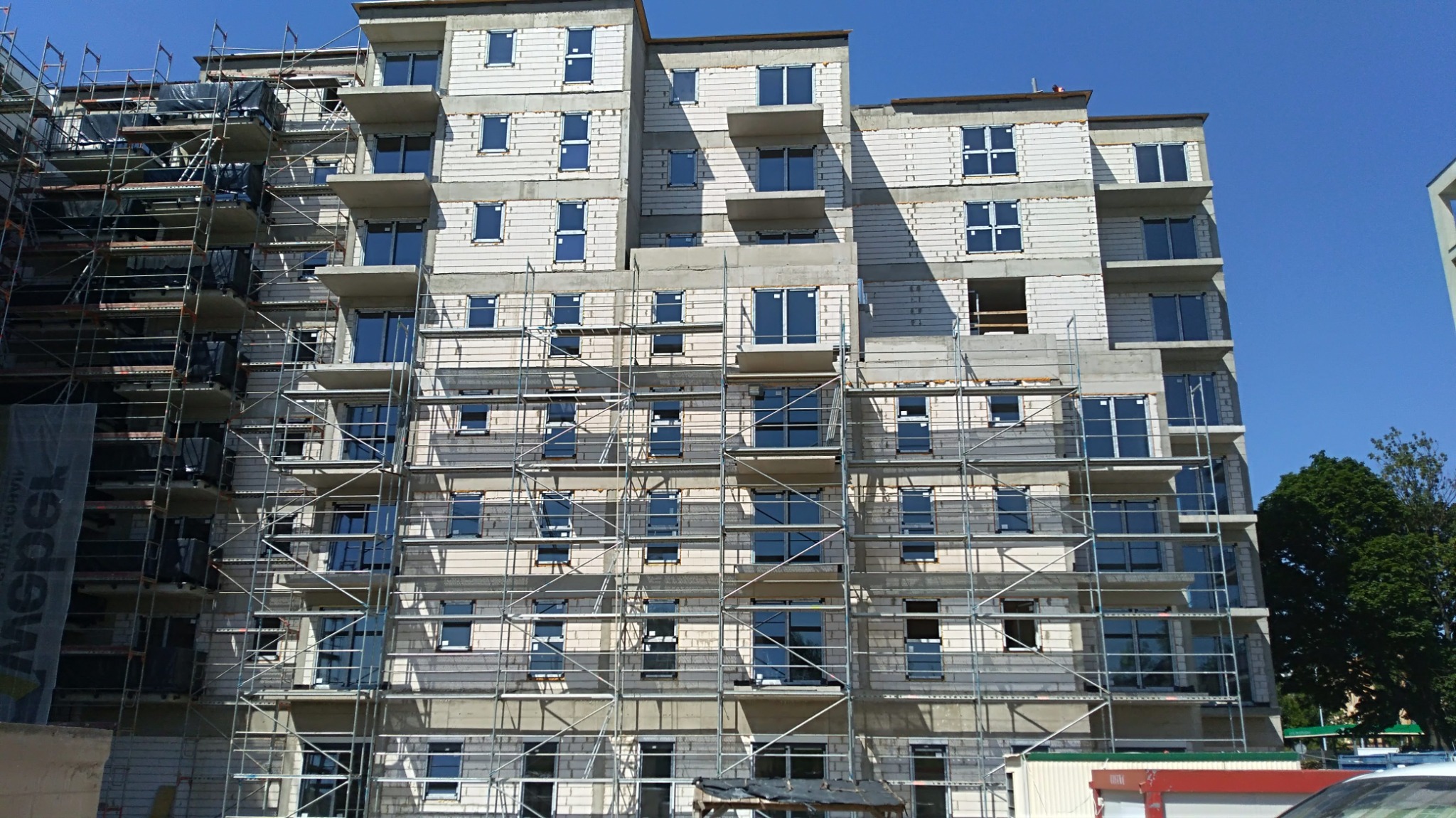 Wielopiętrowy budynek mieszkalny w trakcie budowy, widoczne rusztowania, okna i balkony, ściany z białych bloczków.