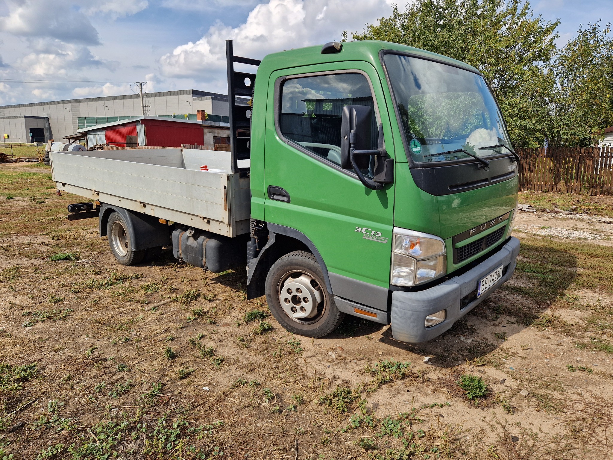 Zielona ciężarówka Fuso Canter z otwartą skrzynią ładunkową, zaparkowana na placu z ubogą roślinnością, w tle budynki przemysłowe i drewniany płot.