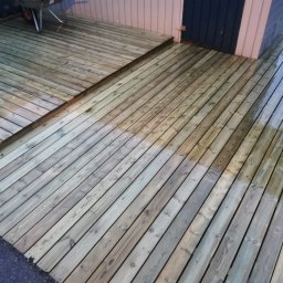 Karol P - Drewniany taras po częściowym czyszczeniu, widoczne różnice w kolorze desek przed i po zabiegu, w tle fragment wózka i ściana budynku.