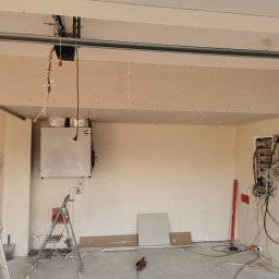 Karol P - Wnętrze garażu w trakcie adaptacji: zamontowana podwieszana konstrukcja z płyt gipsowo-kartonowych, widoczne instalacje wentylacyjne, skrzynka elektryczna z okablowaniem, drabiny i narzędzia.