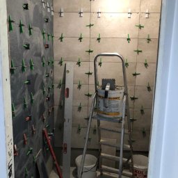 Karol P - Proces układania płytek ściennych w łazience, widoczne poziomica, drabina z wiadrami i klejem, krzyżyki dystansowe w spoinach.