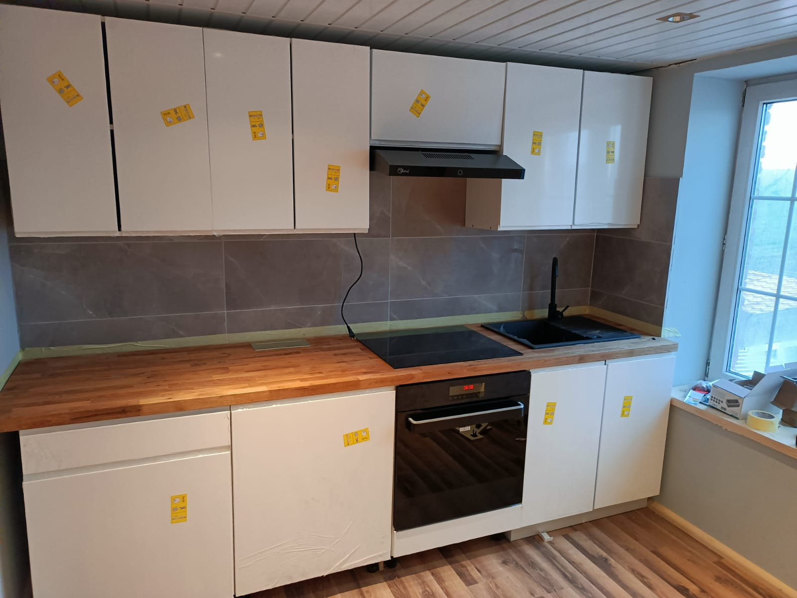 Nowoczesna kuchnia z białymi szafkami, drewnianym blatem i czarnym sprzętem AGD, w trakcie montażu, z folią ochronną na frontach i żółtymi naklejkami.