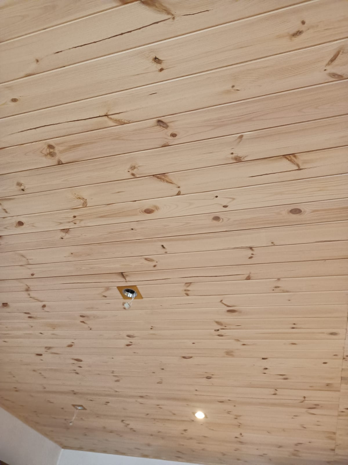 Drewniany sufit z jasnych paneli boazeryjnych z widocznymi sękami i dwoma otworami na oświetlenie, jeden z wystającym kablem.