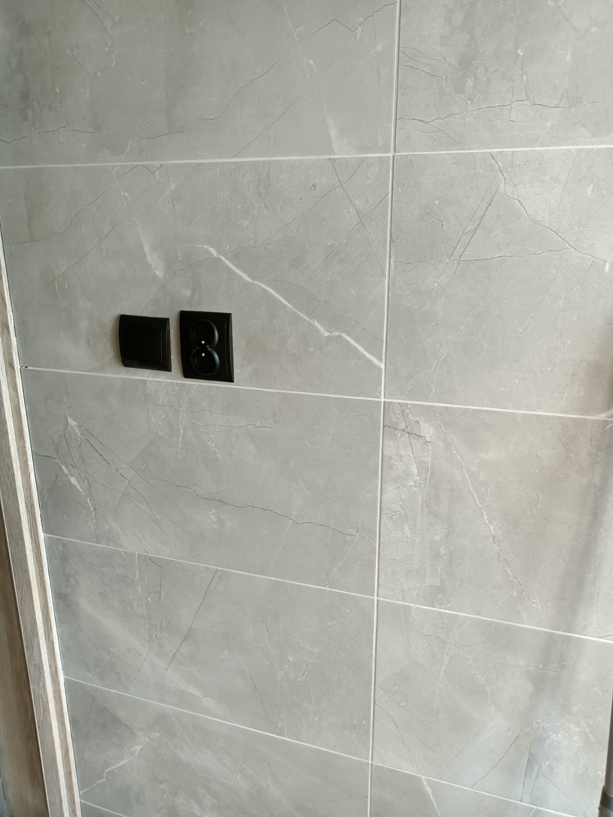 Ściana wyłożona szarymi płytkami imitującymi marmur w Gdańsku, z zamontowanymi czarnymi gniazdkami elektrycznymi i włącznikiem światła, widoczne fugi i fragment ościeżnicy.