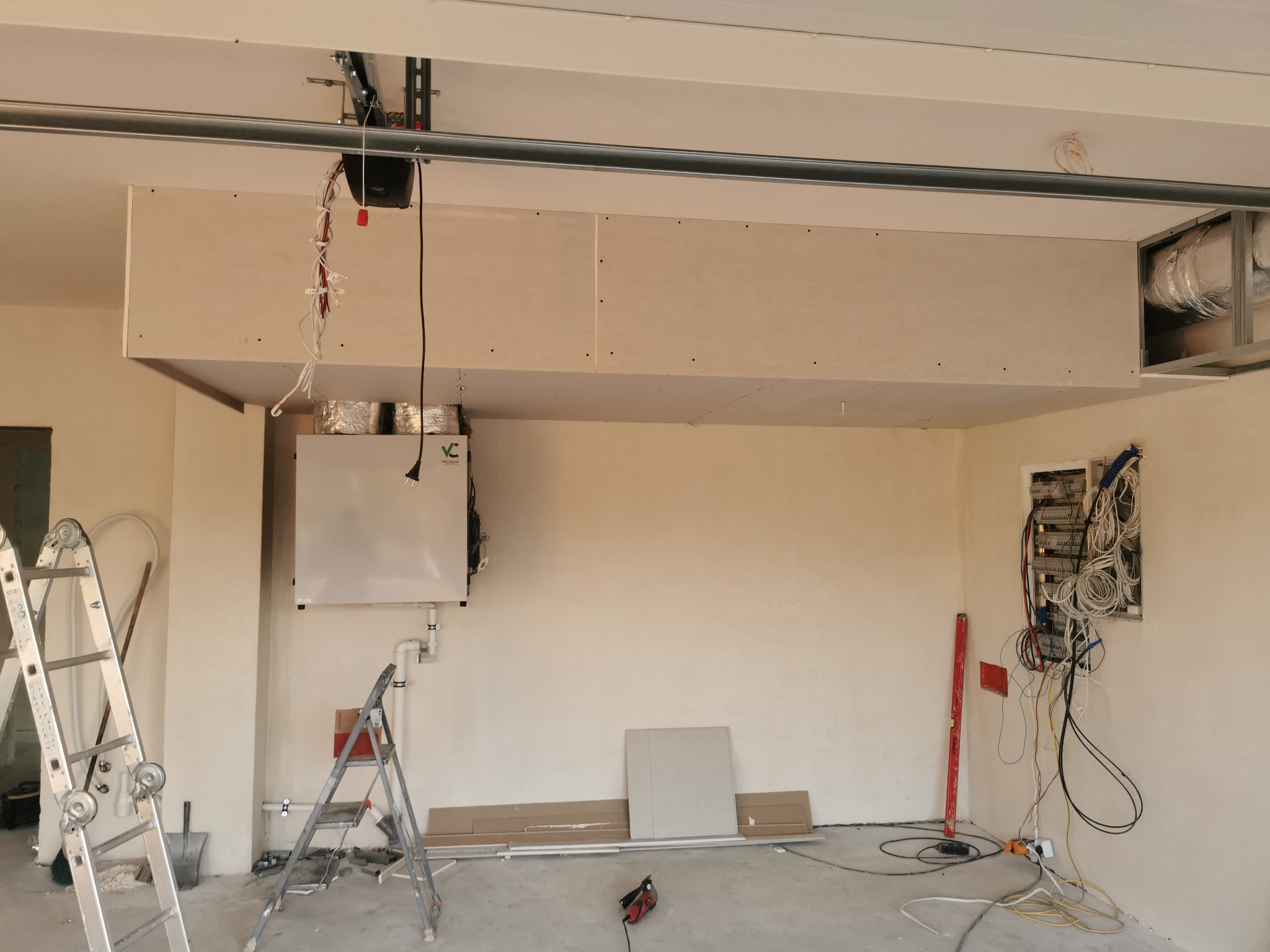 Wnętrze garażu w trakcie adaptacji: zamontowana podwieszana konstrukcja z płyt gipsowo-kartonowych, widoczne instalacje wentylacyjne, skrzynka elektryczna z okablowaniem, drabiny i narzędzia.