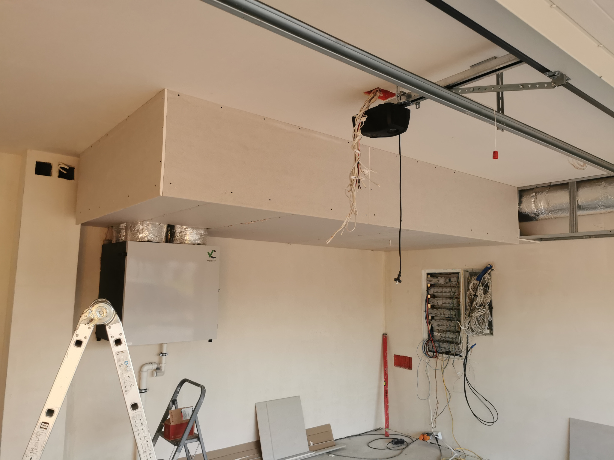 Wnętrze garażu w trakcie adaptacji: zabudowa z płyt gipsowo-kartonowych pod sufitem, widoczny system wentylacyjny z rekuperacją, rozdzielnia elektryczna z okablowaniem, drabina aluminiowa...