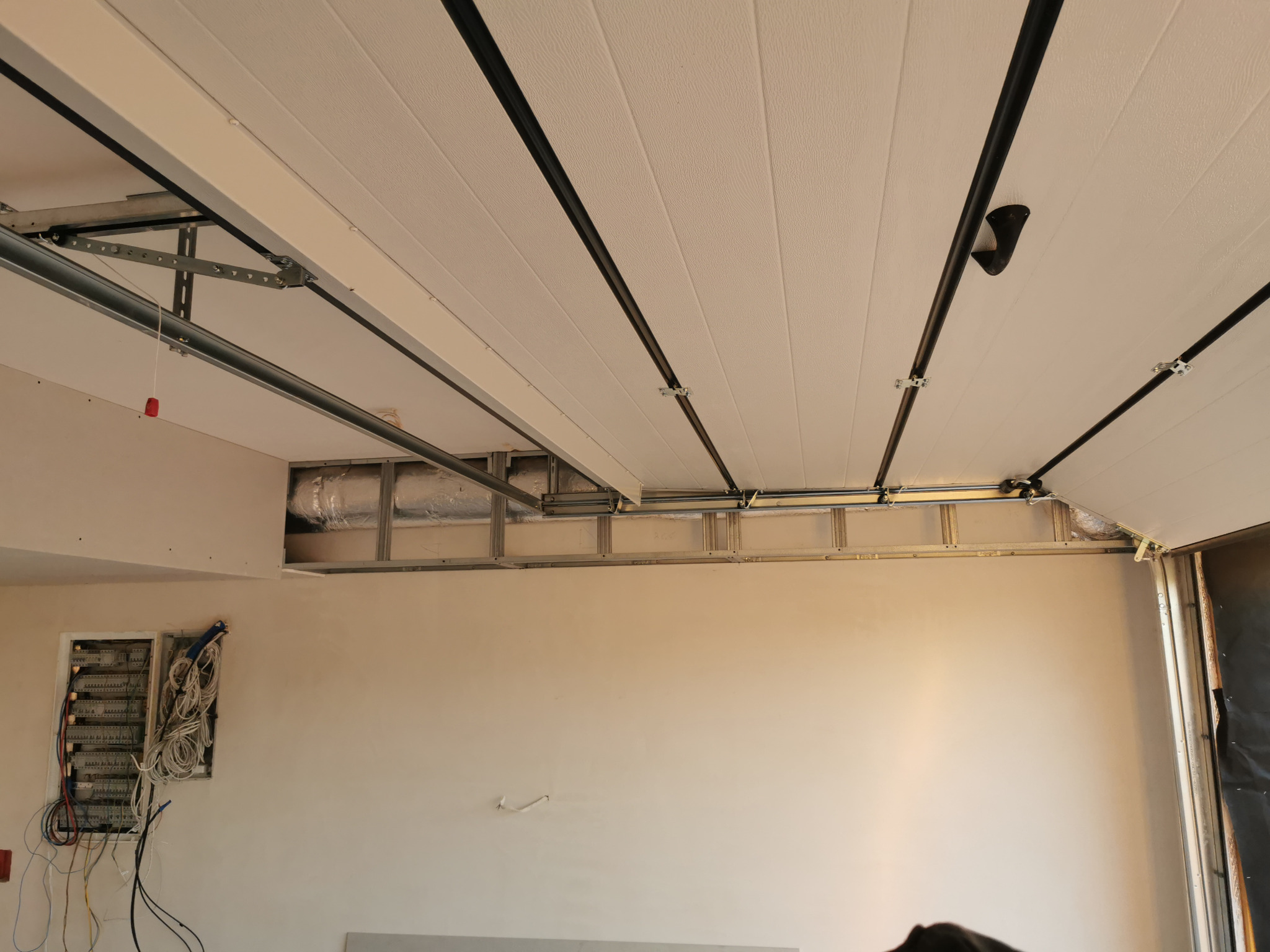 Wnętrze garażu w trakcie wykańczania: widoczna instalacja elektryczna, wentylacja, sufit z paneli z czarnymi listwami i szynami montażowymi.