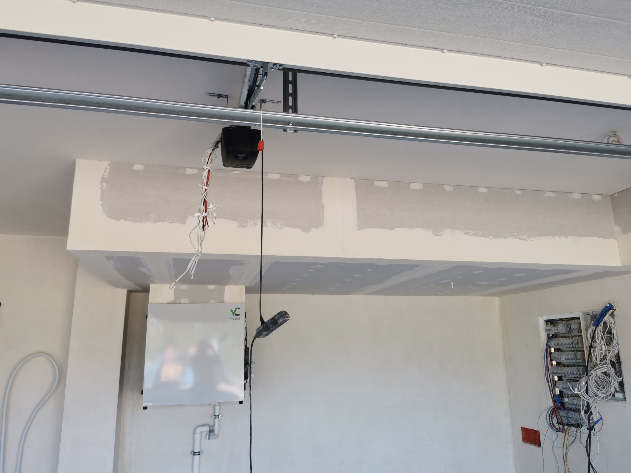 Wnętrze garażu w trakcie wykańczania: widoczny automat do bramy garażowej pod sufitem, fragment ściany z zamontowaną skrzynką elektryczną z okablowaniem oraz centrala wentylacyjna z podłączeniami...