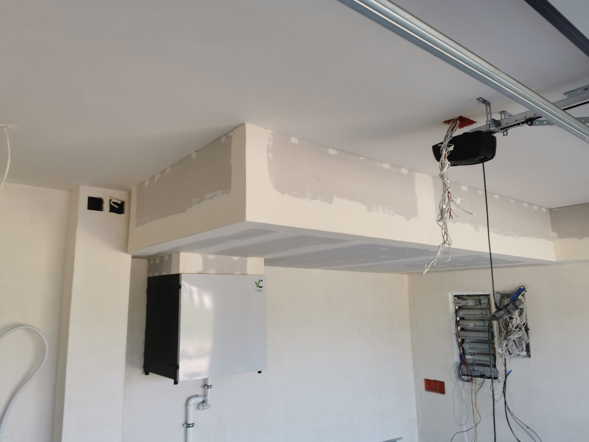 Surowy stan garażu z widoczną instalacją elektryczną, centralą wentylacyjną z rekuperacją i konstrukcją podwieszanego sufitu w trakcie montażu.