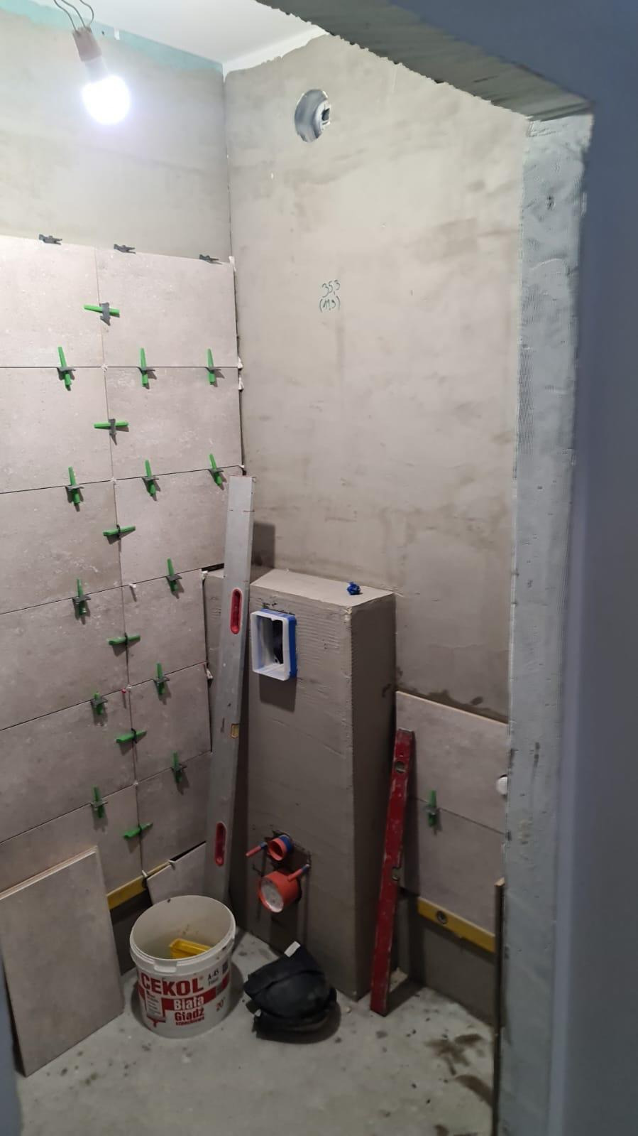 Wnętrze łazienki w trakcie remontu: częściowo ułożone jasne płytki z zielonymi krzyżykami dystansowymi, widoczne poziomice, wiadro z klejem Cekol oraz instalacja wodna w ścianie.