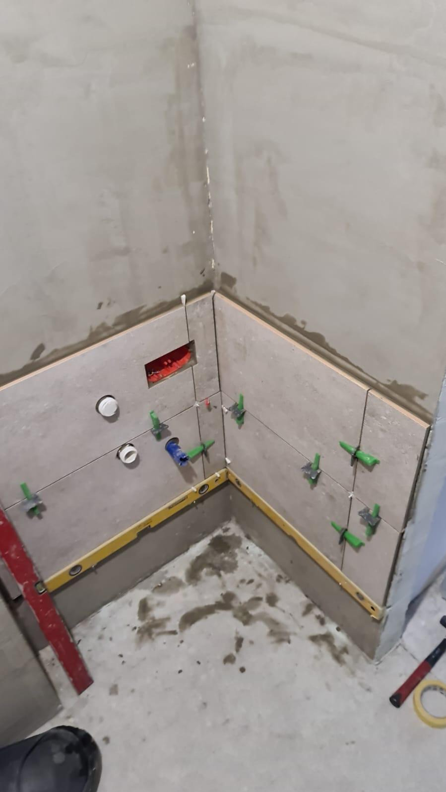 Świeżo ułożone, beżowe płytki ścienne w rogu łazienki, z widocznymi poziomnicami, krzyżykami dystansowymi i otworami na instalację hydrauliczną, w trakcie prac wykończeniowych.