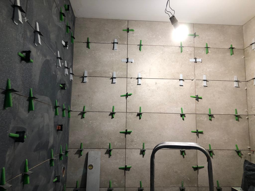 Proces układania beżowych płytek ściennych z użyciem zielonych i białych krzyżyków dystansowych oraz sznurka poziomującego, widoczna drabina i surowy stan ściany po lewej stronie.