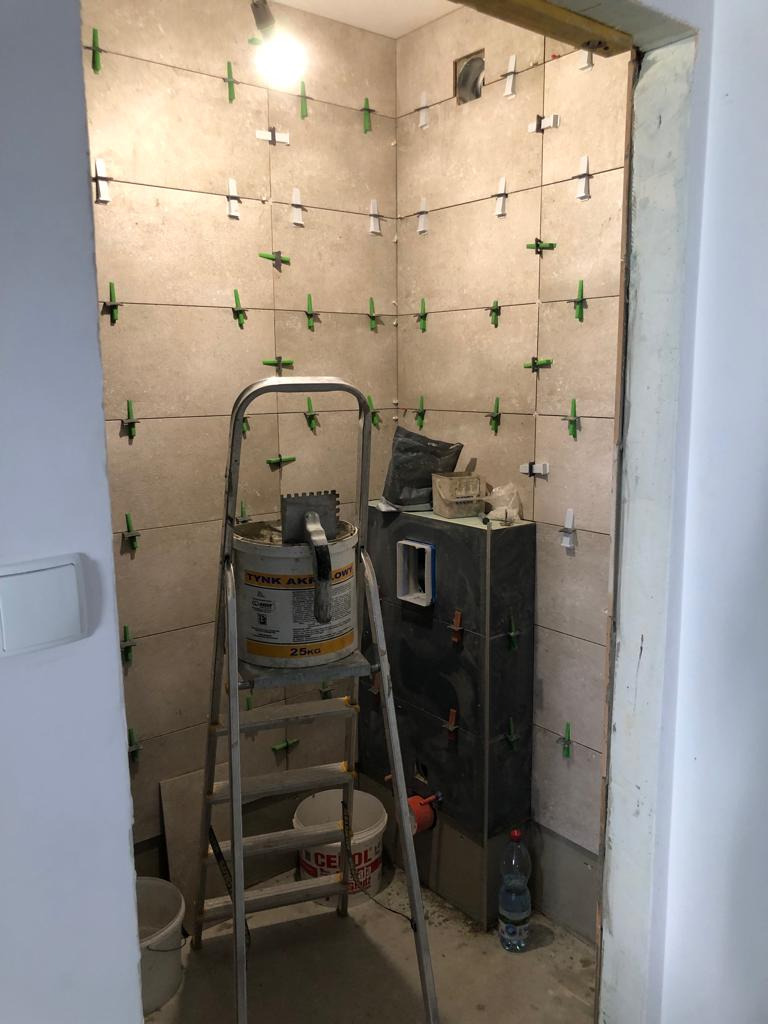 Układanie beżowych płytek w łazience z użyciem poziomicy sznurkowej i zielonych krzyżyków dystansowych, widoczna drabina z wiadrem i puszką kleju oraz zabudowa stelaża WC.