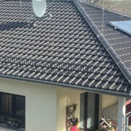 Dom jednorodzinny z brązowym dachem pokrytym dachówką, na którym zamontowana jest antena satelitarna Televes oraz instalacja odgromowa z widocznymi przewodami i odgromnikami. Na dachu widoczny...