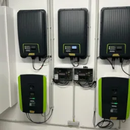 Szereg falowników fotowoltaicznych marki Kostal zamontowanych na białej ścianie, połączonych przewodami w rurkach PCV, z widoczną rozdzielnicą elektryczną po lewej stronie i gniazdkami...